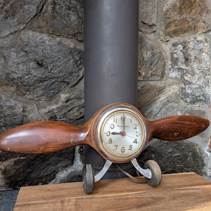 May include: A vintage airplane clock with a wooden propeller and small wheels. The clock face is silver with black numbers and hands. The propeller blades are a dark brown color. The clock is set on a wooden surface.