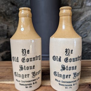 May include: Two vintage stoneware ginger beer bottles. The bottles are beige with a lighter tan top and feature black text that reads "Ye Old Country Stone Ginger Beer" and "Felix Distributors Ltd Vancouver B.C. 284ml."
