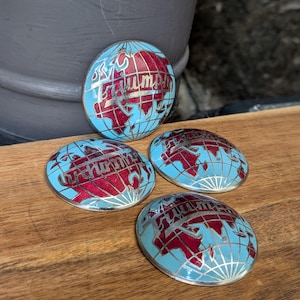 May include: Four oval-shaped Triumph emblems. Each emblem features a light blue background with a red and silver globe design. The word "Triumph" is written in silver script across the globe. The emblems are displayed on a wooden surface.