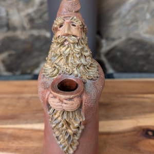 May include: A ceramic figurine of a bearded man wearing a red hat and holding a small brown pot. The figurine is brown with gold accents.