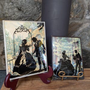 Peut inclure: Deux œuvres d'art en silhouette encadrées représentant des personnages en costume d'époque. L'une est posée sur un support bordeaux, l'autre sur un support doré. Les fonds présentent des scènes extérieures avec des arbres et des éléments architecturaux.