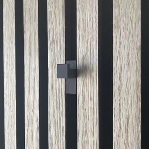Pode incluir: Uma imagem em close-up de um painel de parede com grão de madeira vertical e listras pretas. Uma pequena maçaneta geométrica cinza escuro está presa ao painel. O grão da madeira é claro.