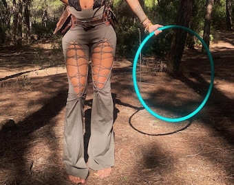 May include: A woman wearing a black halter top with a cut-out design and brown pants with cut-outs. She is holding a turquoise hula hoop in a forest setting.