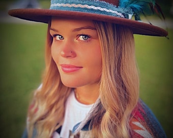 Pode incluir: Uma mulher usando um chapéu fedora de feltro marrom com uma fita azul e branca e uma pena azul. Ela está vestindo uma camisa branca e uma jaqueta estampada colorida.