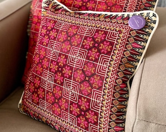 May include: Two decorative square pillows with intricate geometric patterns in shades of red, pink, and gold. The pillows have white piping and are placed on a neutral-toned sofa. A small purple tag is visible on one pillow.
