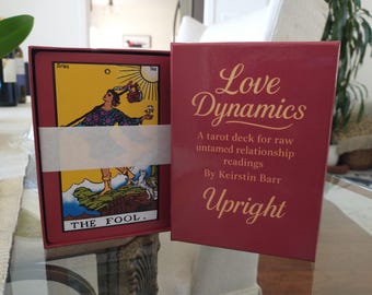 May include: A tarot card deck titled "Love Dynamics" with a burgundy box. The box displays the text "Love Dynamics" in gold script. The deck includes cards with illustrations and the text "THE FOOL."