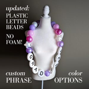 Pode incluir: Um colar de formatura com contas de plástico redondas em tons de roxo, rosa e branco, intercaladas com contas brancas em forma de estrela. Grandes contas circulares brancas exibem o texto preto GRAD 2025. Texto na imagem: updated: PLASTIC LETTER BEADS NO FOAM! custom PHRASE color OPTIONS.
