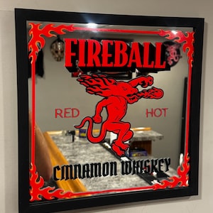 Puede incluir: Espejo cuadrado con marco negro, con el texto rojo "FIREBALL" y un gr&aacute;fico de diablo rojo. Las palabras "RED" y "HOT" flanquean el gr&aacute;fico, y "CINNAMON WHISKEY" est&aacute; en la parte inferior. Un dise&ntilde;o de llama roja bordea el espejo.