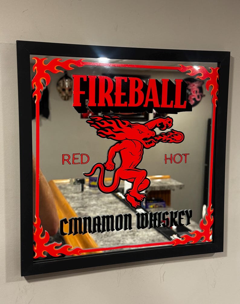 Puede incluir: Espejo cuadrado con marco negro, con el texto rojo "FIREBALL" y un gr&aacute;fico de diablo rojo. Las palabras "RED" y "HOT" flanquean el gr&aacute;fico, y "CINNAMON WHISKEY" est&aacute; en la parte inferior. Un dise&ntilde;o de llama roja bordea el espejo.