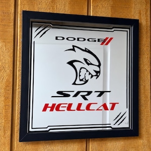 May include: A square mirror in a black frame featuring the Dodge logo, the SRT Hellcat cat head emblem, and the text SRT HELLCAT in black and red. The design includes stylized claw marks in the top left and bottom right corners of the mirror surface.