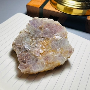 May include: A raw, natural fluorite crystal with a mix of colors, including purple, yellow, and clear. The crystal is rough and textured, with visible crystalline structures. It sits on a lined notepad.