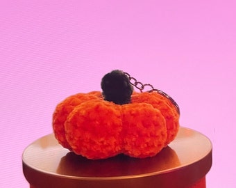 Puede incluir: Una pequeña calabaza de peluche naranja con un tallo negro y una cadena plateada. La calabaza está sobre una superficie dorada, sobre un fondo rosa. Este artículo decorativo es perfecto para el otoño y Halloween.