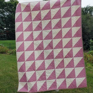 May include: A large, rectangular quilt featuring a repeating pattern of pink and cream-coloured triangles. The quilt is displayed outdoors, with a green lawn and trees in the background. The triangles are arranged in rows and columns, creating a geometric design.