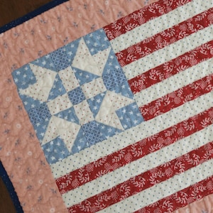 May include: A rectangular quilted wall hanging featuring a stylized American flag design. The canton displays a blue and cream star pattern, while the stripes alternate between red floral fabric and cream fabric. It has a peach border and blue trim.