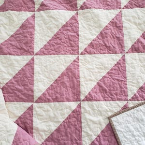 May include: A handmade quilt with a repeating pattern of pink triangles on a white background. The quilt has a quilted texture and a brown border. The design creates a geometric pattern, suitable for a bedspread or decorative throw.
