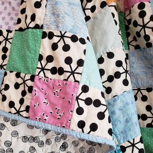 May include: A patchwork quilt with a variety of patterns and colors. The quilt features squares with black and white geometric designs, pink and green floral patterns, and blue butterfly and feather motifs. The quilt's edge is trimmed with a light blue dotted fabric.