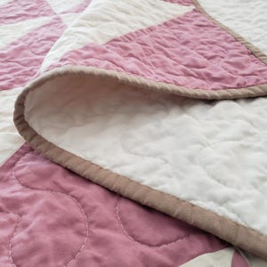 May include: A close-up of a handmade quilt with a starburst pattern. The quilt features alternating sections of white and dusty rose fabric, with a brown border. The stitching is visible, adding texture to the design.