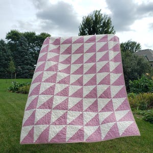 May include: A large, handmade quilt featuring a repeating pattern of pink and white triangles. The quilt is displayed outdoors, with a green lawn and trees in the background. The quilt's design creates a geometric pattern. The quilt is likely to be approximately 200cm x 200cm.