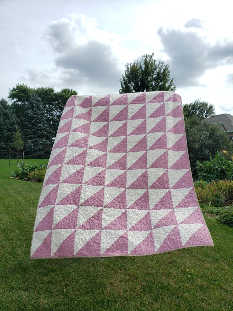 May include: A large, handmade quilt featuring a repeating pattern of pink and white triangles. The quilt is displayed outdoors, with a green lawn and trees in the background. The quilt's design creates a geometric pattern. The quilt is likely to be approximately 200cm x 200cm.