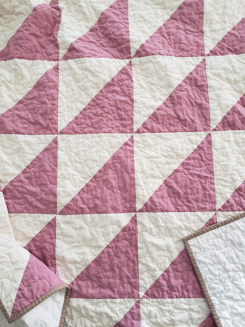 May include: A handmade quilt with a repeating pattern of pink triangles on a white background. The quilt has a quilted texture and a brown border. The design creates a geometric pattern, suitable for a bedspread or decorative throw.