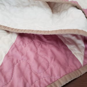 May include: Close-up of a quilted blanket with a patchwork design. The blanket features sections of white and pink fabric, with a light brown border. The stitching is visible, and the blanket appears soft and comfortable.