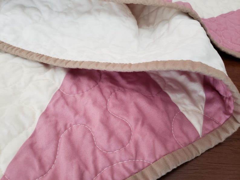 May include: Close-up of a quilted blanket with a patchwork design. The blanket features sections of white and pink fabric, with a light brown border. The stitching is visible, and the blanket appears soft and comfortable.