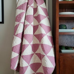 May include: A pink and white patterned quilt, featuring a design of curved triangles. The quilt is draped over a white ladder, highlighting its texture and pattern. The soft colours and design create a cosy aesthetic. The quilt is approximately 150cm x 200cm.