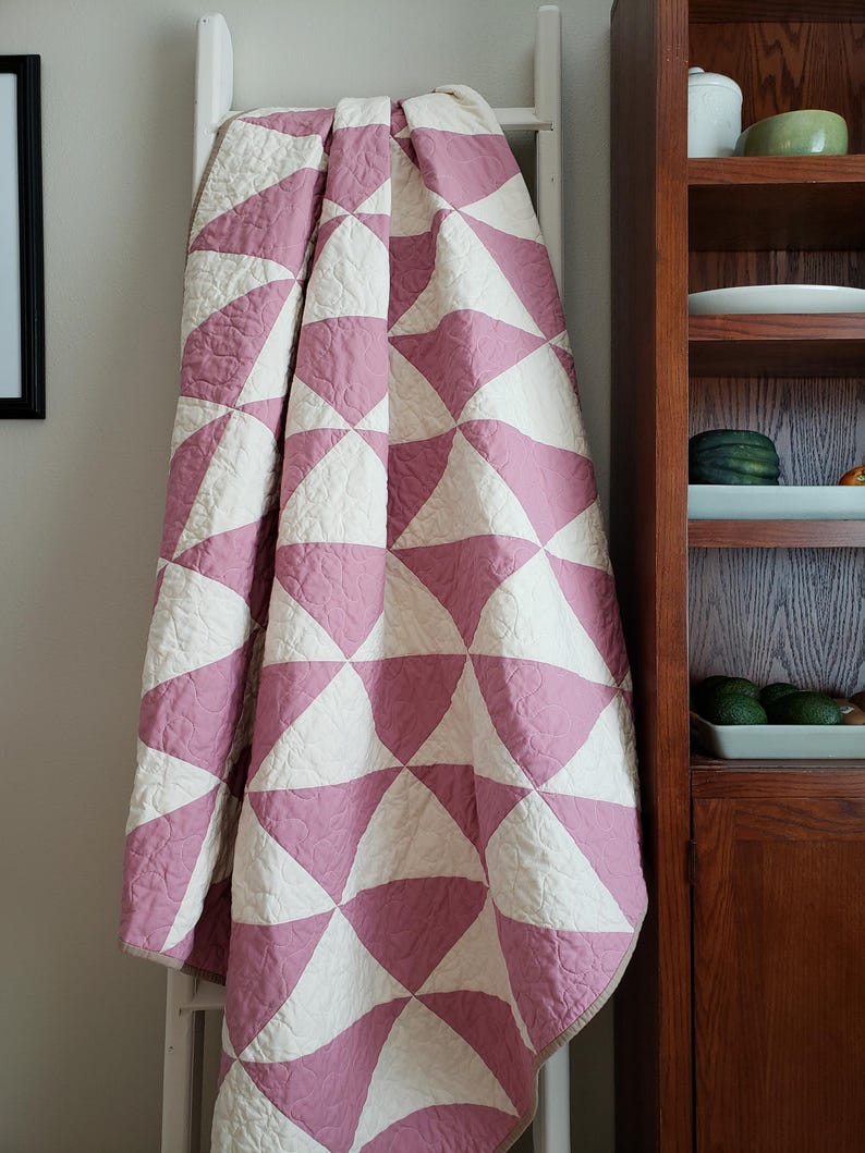 May include: A pink and white patterned quilt, featuring a design of curved triangles. The quilt is draped over a white ladder, highlighting its texture and pattern. The soft colours and design create a cosy aesthetic. The quilt is approximately 150cm x 200cm.