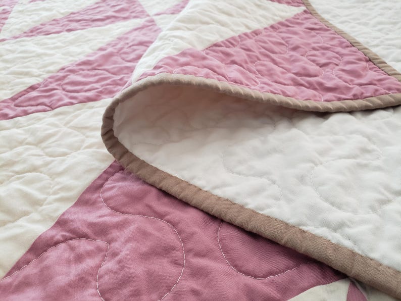 May include: A close-up of a handmade quilt with a starburst pattern. The quilt features alternating sections of white and dusty rose fabric, with a brown border. The stitching is visible, adding texture to the design.