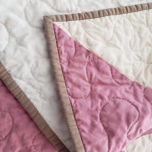 May include: Close-up of a quilted blanket with a floral pattern. The blanket features white and dusty rose pink fabric, with a brown border. The stitching creates a textured, soft appearance. The blanket is folded, revealing the contrasting colours.
