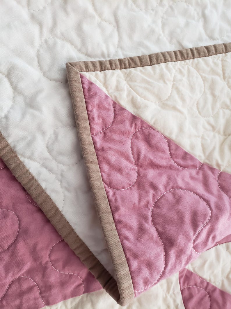 May include: Close-up of a quilted blanket with a floral pattern. The blanket features white and dusty rose pink fabric, with a brown border. The stitching creates a textured, soft appearance. The blanket is folded, revealing the contrasting colours.
