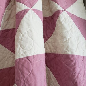 May include: A close-up of a folded quilt with a starburst pattern. The quilt features alternating sections of white and mauve fabric. The fabric is quilted with a wavy stitch pattern. The quilt is made of soft, quilted fabric.