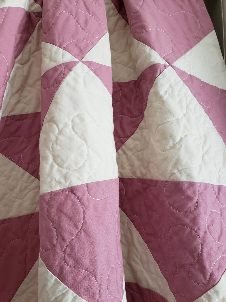 May include: A close-up of a folded quilt with a starburst pattern. The quilt features alternating sections of white and mauve fabric. The fabric is quilted with a wavy stitch pattern. The quilt is made of soft, quilted fabric.