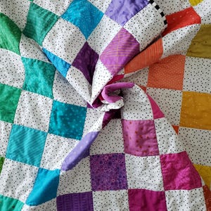 May include: A patchwork quilt featuring a grid of multicolored fabric squares separated by white fabric with small black dots. The squares include shades of green, blue, purple, pink, orange, and yellow, with various subtle patterns on each square.