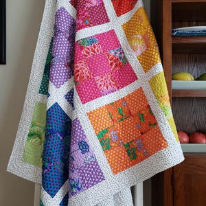 May include: A patchwork quilt featuring a grid of colorful fabric squares in shades of purple, blue, green, orange, pink, and yellow, separated by white sashing with small black dots. The quilt displays various floral and polka dot patterns.