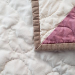 May include: Close-up of a quilted blanket with a soft, textured surface. The quilt features a pattern of stitched circles on a cream-coloured fabric. A section of pink fabric is visible, bordered by a brown edge.