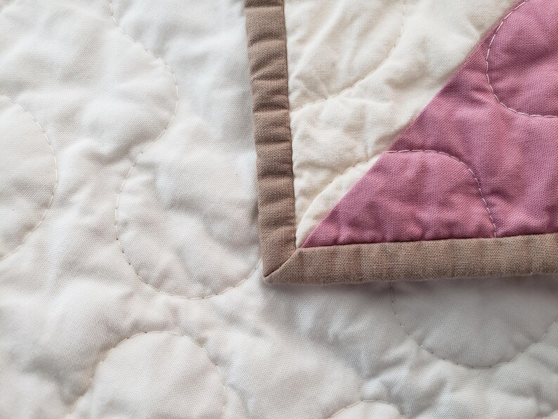 May include: Close-up of a quilted blanket with a soft, textured surface. The quilt features a pattern of stitched circles on a cream-coloured fabric. A section of pink fabric is visible, bordered by a brown edge.