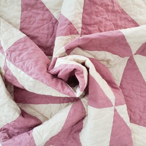 May include: Close-up of a folded quilt with a geometric pattern. The quilt has alternating sections of white and pink fabric, with a quilted design. The fabric is soft and appears to be cotton.