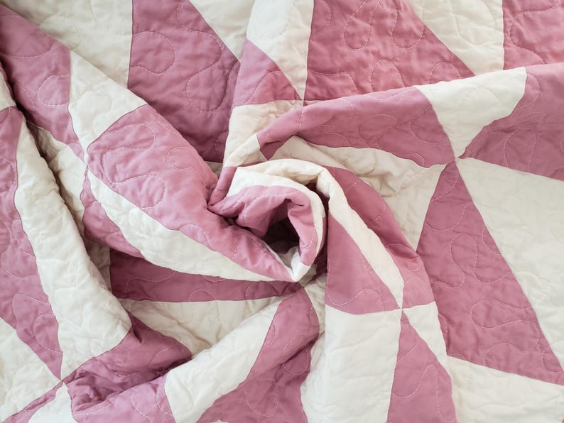 May include: Close-up of a folded quilt with a geometric pattern. The quilt has alternating sections of white and pink fabric, with a quilted design. The fabric is soft and appears to be cotton.