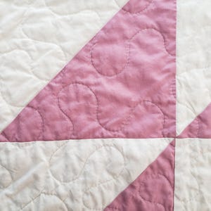May include: Close-up of a quilted textile with a repeating pattern of white and pink squares and triangles. The pink sections have a mottled appearance and are stitched with a curved design. The quilt is a handmade item.