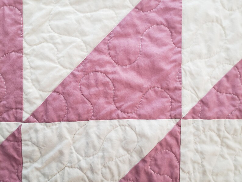 May include: Close-up of a quilted textile with a repeating pattern of white and pink squares and triangles. The pink sections have a mottled appearance and are stitched with a curved design. The quilt is a handmade item.