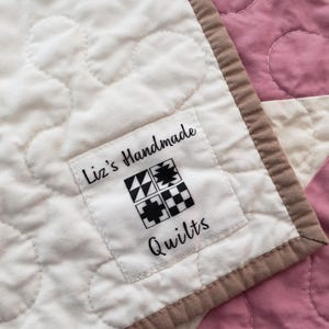 May include: A close-up shot of a handmade quilt with a white background and pink sections. The quilt has a label that says "Liz's Handmade Quilts" with a geometric design. It has a brown border.