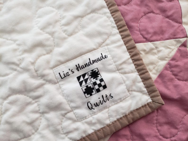 May include: A close-up shot of a handmade quilt with a white background and pink sections. The quilt has a label that says "Liz's Handmade Quilts" with a geometric design. It has a brown border.