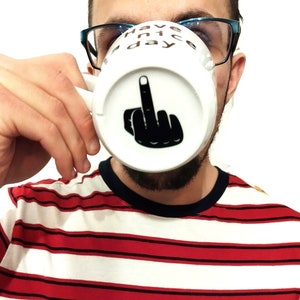 Peut inclure: Une tasse en céramique blanche avec un motif noir représentant une main faisant un geste du majeur sur le dessous extérieur. Le texte "Have a nice day" est imprimé en lettres noires près du bord supérieur de la tasse, munie d'une anse.