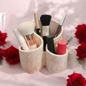 Puede incluir: Un organizador de maquillaje de cerámica marmoleado en blanco y rosa con cuatro compartimentos. El organizador contiene varios pinceles de maquillaje, un lápiz labial y un delineador de ojos negro.