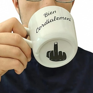 Puede incluir: Una taza de cerámica blanca con el texto negro Bien Cordialement impreso en la parte superior y una ilustración gráfica negra de una mano haciendo un gesto de dedo medio en la parte inferior de la superficie exterior de la taza.