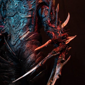 Könnte beinhalten: Nahaufnahme eines detaillierten, strukturierten Kostüms mit aufwendiger Panzerung. Das Kostüm weist scharfe, klauenartige Verlängerungen an Arm und Schulter auf, mit einer Farbpalette aus Schwarz, Rot und Blau. Das Design deutet auf ein Fantasy- oder Sci-Fi-Thema hin.