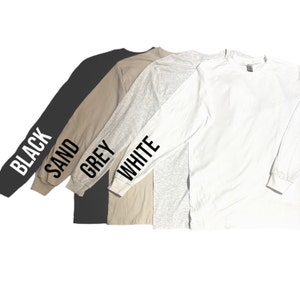Puede incluir: Cuatro camisas de manga larga en negro, arena, gris y blanco. Cada camisa tiene el nombre del color impreso en la manga en negro.