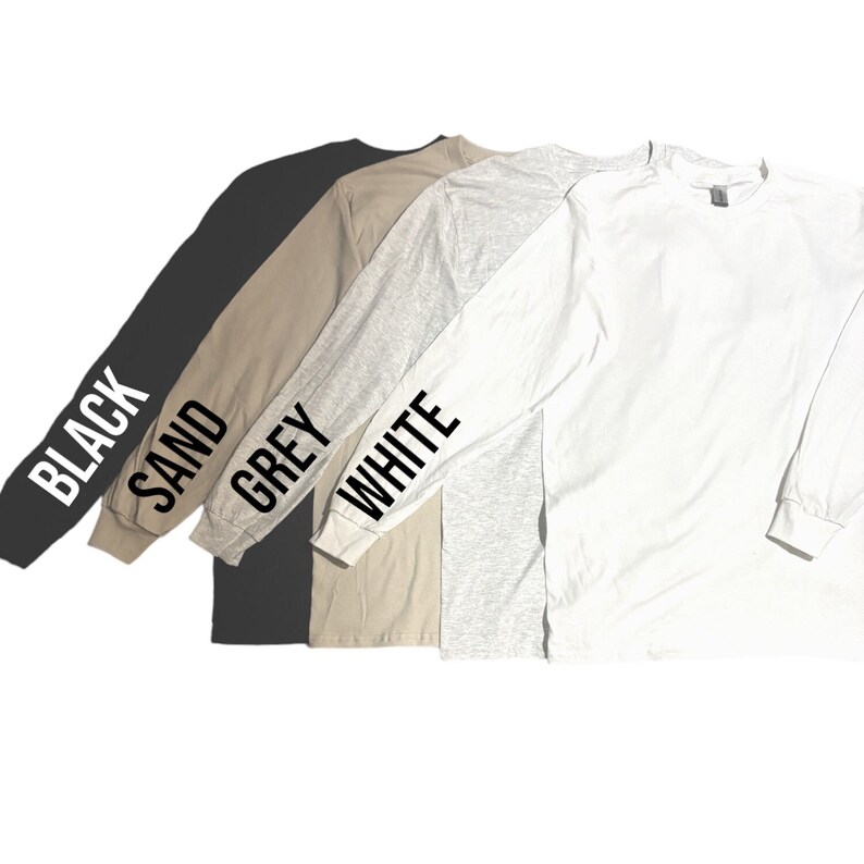Puede incluir: Cuatro camisas de manga larga en negro, arena, gris y blanco. Cada camisa tiene el nombre del color impreso en la manga en negro.