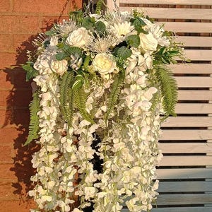 May include: A white hanging flower basket with white flowers and green ferns. The basket is hanging from a black metal hook on a brick wall.